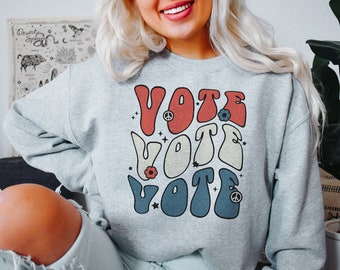 May include: A heather gray crewneck sweatshirt featuring a graphic print with the word VOTE repeated three times in a stacked, wavy font. The text colors are red, cream, and blue, accented with small star, flower, and peace sign symbols.