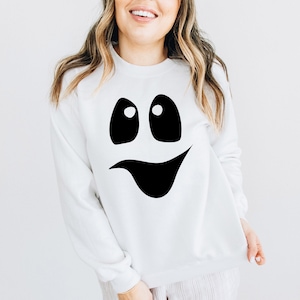 May include: White sweatshirt featuring a large black ghost face design. The ghost face has two oval eyes and a curved mouth. The sweatshirt is a crew neck style and is made of a soft material. This is a Halloween-themed apparel item.