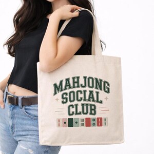 Puede incluir: Un bolso de tela beige con el texto verde oscuro "MAHJONG SOCIAL CLUB" en una fuente universitaria. Debajo del texto hay pequeñas ilustraciones gráficas de fichas de mahjong en tonos verde, rojo y beige, acentuadas por pequeñas estrellas.
