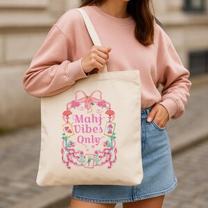 Puede incluir: Un bolso de tela color crema con un estampado gráfico en rosa y multicolor. El diseño incluye un lazo rosa grande, elementos florales y fichas de mahjong dispuestas en un marco. El texto "Mahj Vibes Only" está impreso en letras rosas.