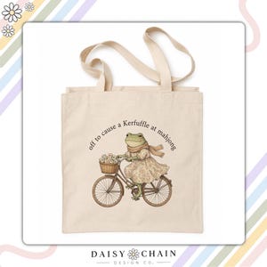 Puede incluir: Una bolsa de tela beige con una ilustración impresa de una rana con vestido y bufanda, montada en una bicicleta con una cesta llena de fichas de mahjong. El texto sobre la rana dice, off to cause a Kerfuffle at mahjong.