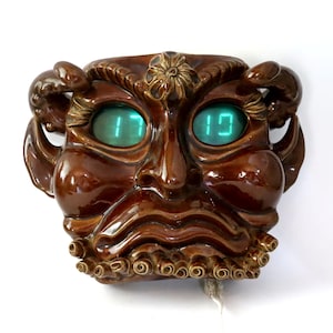 May include: A brown ceramic wall clock shaped like a stylized face with green digital eyes displaying the time 17:10. The clock has ornate details, including a floral accent and curly embellishments.