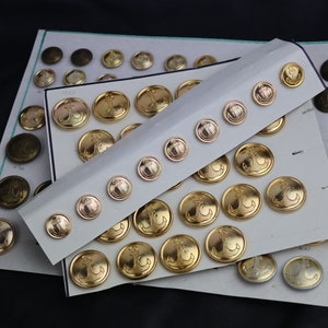 Può includere: Una collezione di bottoni in metallo color oro con un design a forma di ancora. I bottoni sono disposti su cartoncini bianchi, con alcuni bottoni su un cartoncino marrone.