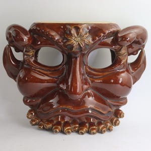 May include: A glossy, brown ceramic mask with a detailed, expressive face. The mask features large eye openings, a prominent nose, and a detailed mouth area. The design includes ear-like protrusions and a floral accent.