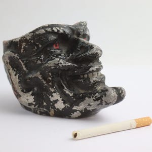 Könnte beinhalten: Ein Vintage-Aschenbecher in Form eines grotesken Gesichts mit roten Augen, schwarz mit silbernen Akzenten bemalt. Eine brennende Zigarette liegt daneben. Der Aschenbecher hat eine breite Öffnung für Asche.