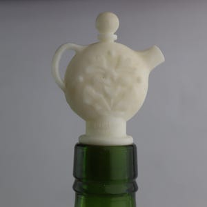 May include: White ceramic bottle stopper shaped like a teapot with floral details. The stopper is on top of a green glass bottle.