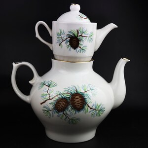 Könnte beinhalten: Eine weiße Porzellan-Teekanne mit einem Design aus Tannenzapfen und -zweigen. Die Teekanne hat eine kleinere Teekanne oben drauf, ebenfalls mit einem Design aus Tannenzapfen und -zweigen.