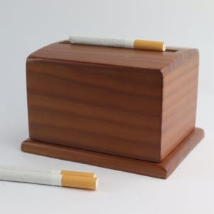 Pode incluir: Uma caixa de madeira com tampa articulada e uma ranhura para cigarros. Dois cigarros estão sobre uma superfície branca ao lado da caixa.
