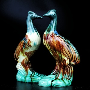 May include: Two ceramic bird figurines facing each other. The birds are painted in shades of brown, green, and blue with a mottled glaze. They are standing on a base that is also painted in shades of green and blue.