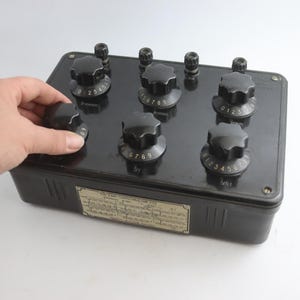 Könnte beinhalten: Ein schwarzes elektronisches Bauteil mit mehreren nummerierten Knöpfen. Eine Hand dreht einen der Knöpfe. Das rechteckige Bauteil hat ein Etikett mit Text. Die Knöpfe sind von 0 bis 9 nummeriert.