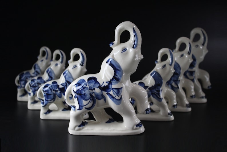 May include: A collection of white ceramic elephant figurines with blue floral designs. The elephants are arranged in a row, each with its trunk raised. The figurines are on a dark surface.