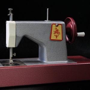 May include: A vintage toy sewing machine with a red base and a gray metal body. The machine has a white plastic top and a small floral design on the side.