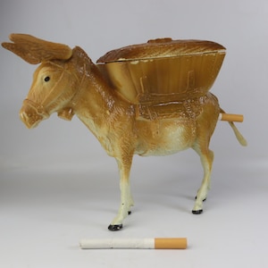 Pode incluir: Um cinzeiro de novidade em forma de burro carregando uma cesta nas costas. O burro é bege com detalhes brancos nas pernas e cascos pretos. Um cigarro aceso está na cauda do burro. Um cigarro está na superfície.