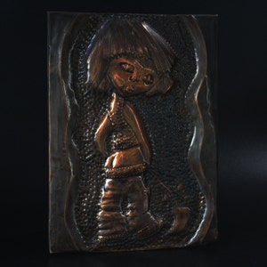 May include: A copper wall sculpture of a stylized figure with a short haircut and a textured surface. The figure is wearing a long-sleeved shirt and pants.