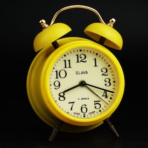 May include: A yellow alarm clock with black numbers and hands. The clock face reads "Slava" and "11 Jewels" and "Made in USSR".