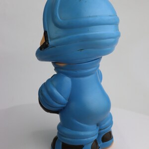 K&ouml;nnte beinhalten: Eine blaue Plastikspielzeug-Astronautenfigur mit Helm und schwarzen Stiefeln. Die Figur steht auf einer wei&szlig;en Oberfl&auml;che.