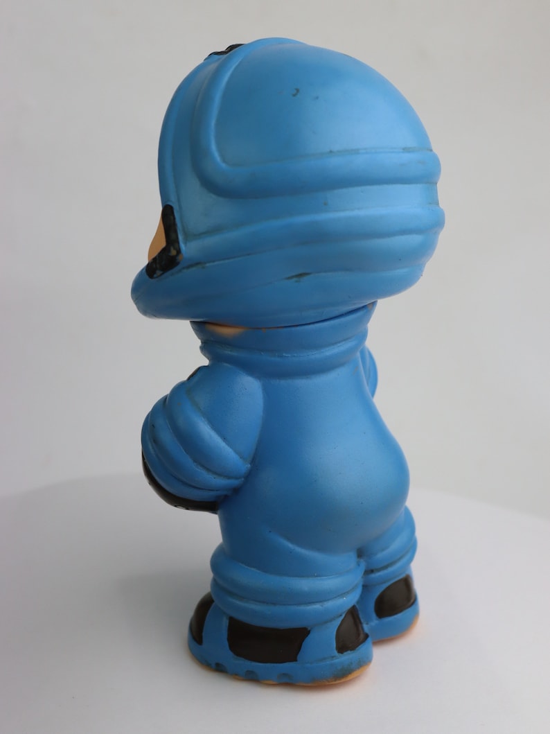 K&ouml;nnte beinhalten: Eine blaue Plastikspielzeug-Astronautenfigur mit Helm und schwarzen Stiefeln. Die Figur steht auf einer wei&szlig;en Oberfl&auml;che.
