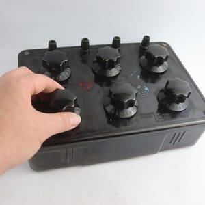 Puede incluir: Un componente eléctrico negro con seis perillas negras, cada una numerada del 0 al 5. El componente es rectangular con ranuras de ventilación en el lateral. Una mano está ajustando una de las perillas.