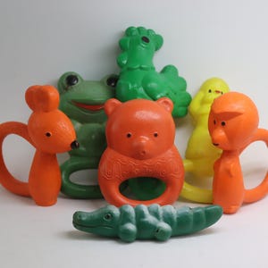 May include: A collection of colorful vintage baby toys. The toys include a green frog, a green alligator, a yellow duck, and orange bear, rabbit, and monkey-shaped teethers. The toys are made of plastic and have a ring for easy gripping.