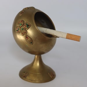 Może przedstawiać: Zabytkowa mosiężna popielniczka z misą w kształcie jaja i podstawą na cokole. Popielniczka posiada kolorowe inkrustowane wzory i zawiera zapalonego papierosa. Mosiądz ma ciepły, złoty odcień.