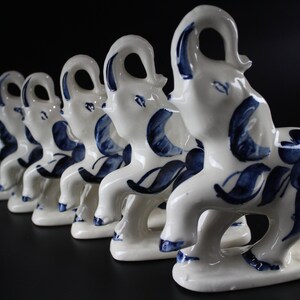 May include: A row of white ceramic elephant figurines with blue floral patterns. The elephants are standing on a base and have their trunks raised. The background is black.