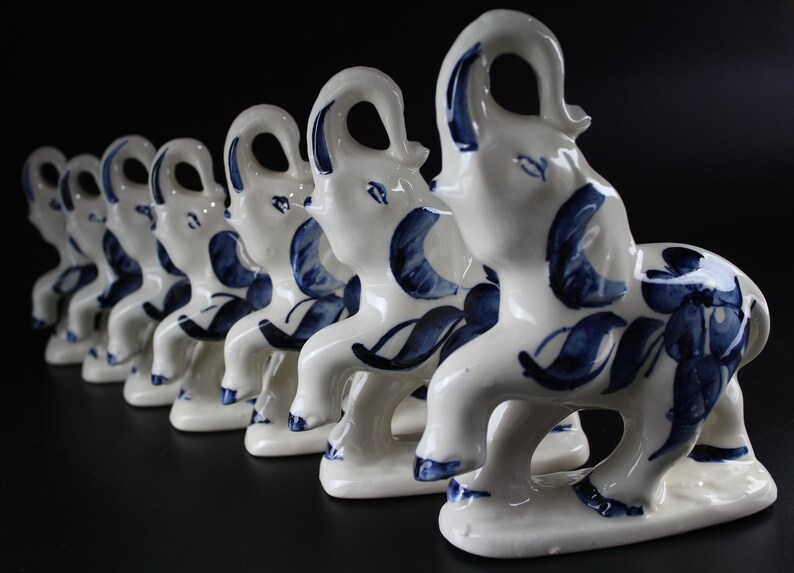 May include: A row of white ceramic elephant figurines with blue floral patterns. The elephants are standing on a base and have their trunks raised. The background is black.