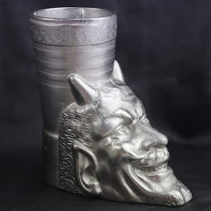 May include: A silver metal cup shaped like a devil's head. The cup has a cylindrical top with a decorative band around the rim.
