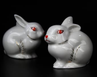 May include: Two white porcelain rabbit figurines with red eyes and gold accents. The rabbits are sitting on a black surface.