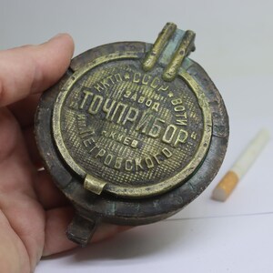 May include: Vintage round metal ashtray with the text "TOCHPRYBOR" in Cyrillic script. The ashtray is a tarnished bronze color and has a hinged lid. A cigarette rests nearby.