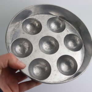 May include: A vintage, round, aluminum escargot dish with seven individual compartments. The dish has a slightly worn, metallic finish, suggesting age and use. The circular design is ideal for serving snails or other small appetizers.