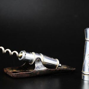 May include: A silver corkscrew with a metal base and a separate silver stopper. The corkscrew is shaped like a cannon with a spiral corkscrew attached to the barrel.
