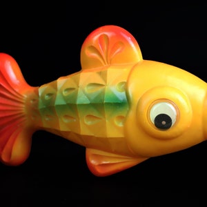 May include: A yellow, orange, and green plastic toy fish with a large, round, white eye. The fish has a red, orange, and yellow tail fin.