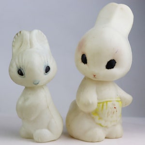 May include: Two vintage white rubber bunny figurines. One bunny is standing upright, the other is sitting. The bunnies have black eyes and are wearing yellow aprons. The figurines are likely from the mid-20th century.