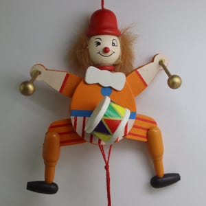 May include: A wooden pull toy clown with a red hat, orange and white striped pants, and a colorful drum. The clown has a winking eye and a big smile.