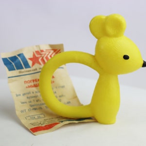 May include: A bright yellow, vintage-style mouse-shaped baby toy with a circular handle. The toy has black eyes and a black nose. A piece of aged paper with text and a red star is behind the toy.