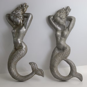 May include: Two silver-colored mermaid wall sculptures. Each mermaid has long flowing hair, arms raised behind the head, and a fishtail. The sculptures are made of a metallic material and are displayed against a white background.