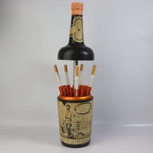 May include: Vintage cigarette holder with a black bottle-shaped top and a cylindrical base. The base features a sepia-toned illustration of a man. Cigarettes are visible inside the holder.