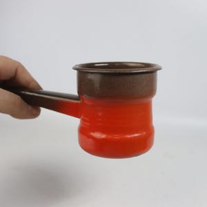 May include: A small, vintage, red and brown enamelware coffee pot with a handle. The pot has a rounded body and a dark brown rim. The handle is also brown. The pot is held against a white background.