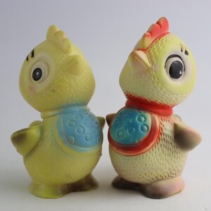 May include: Two yellow rubber toy chickens with blue and red accents. One chicken has a blue bib with white polka dots and the other has a red bib with blue polka dots.