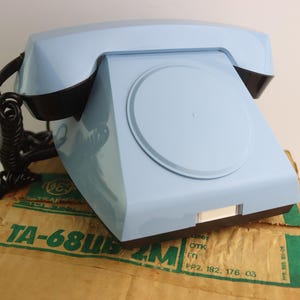 Puede incluir: Un teléfono de disco rotatorio azul claro con un auricular negro y un cable en espiral. El teléfono está sentado en una caja de cartón marrón con texto verde que dice "TA-68ШБ 2M ПП2. 182. 176-03".