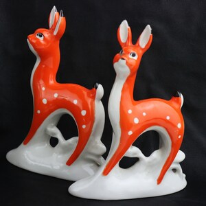 Op de afbeelding: Twee porseleinen hertenfiguren met witte bases en oranje lichamen met witte vlekken. De herten staan op een witte basis met een gestileerde tak.