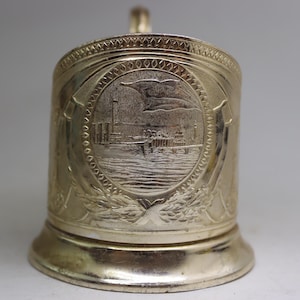 May include: Silver-toned metal cup holder with an engraved design of a cityscape and a ship. The cup holder has a decorative rim and a base.