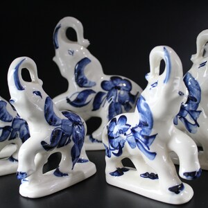 May include: A collection of white ceramic elephant figurines with blue floral and leaf designs. The elephants have their trunks raised and are arranged on a dark surface. The figurines are decorative.