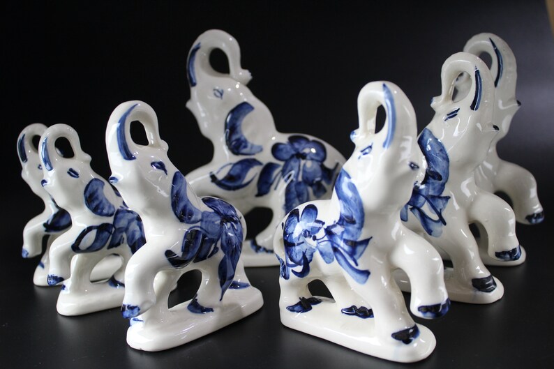 May include: A collection of white ceramic elephant figurines with blue floral and leaf designs. The elephants have their trunks raised and are arranged on a dark surface. The figurines are decorative.