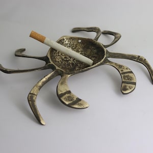 May include: A vintage brass ashtray shaped like a crab, with a cigarette resting in the center. The ashtray has six legs and a textured surface, showing signs of age and use. The cigarette is white with a brown filter.