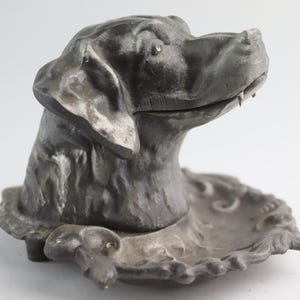 Peut inclure: Un cendrier en métal argenté vintage en forme de tête de chien. La tête du chien est sculptée dans un style réaliste avec un museau et des oreilles détaillés. Le cendrier est une pièce décorative avec un bord festonné.