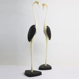 May include: Two white wooden bird sculptures with black heads and long necks. The birds are facing each other and are standing on black bases.