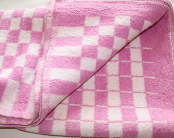 Peut inclure: Une couverture à carreaux rose et blanc avec une texture douce et moelleuse. La couverture est pliée en deux et le motif à carreaux est visible des deux côtés.