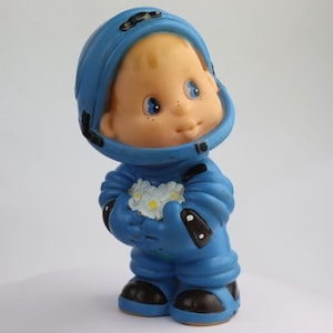 K&ouml;nnte beinhalten: Ein blaues Astronautenspielzeug aus Gummi, das einen Strau&szlig; wei&szlig;er Blumen h&auml;lt. Das Spielzeug hat einen gro&szlig;en Kopf mit einem blauen Helm und ein l&auml;chelndes Gesicht.