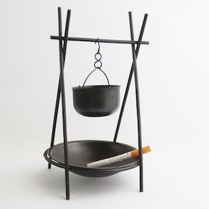 May include: A black metal ashtray with a hanging pot and a cigarette. The ashtray has a tripod base and a circular bowl. The pot hangs from a metal frame. The cigarette is resting in the bowl.